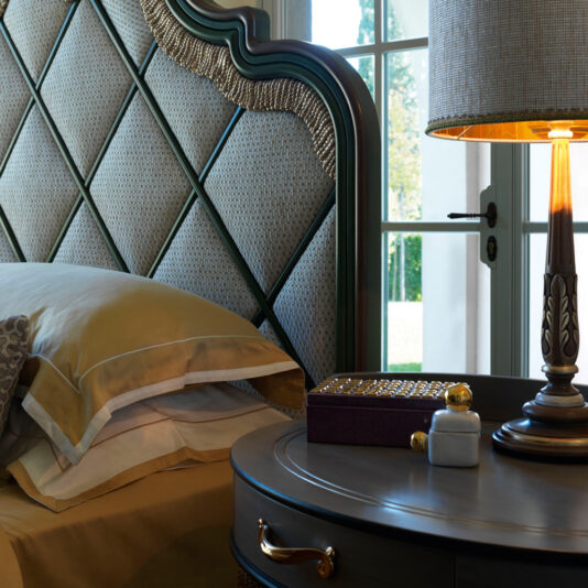 Exclusive Bedside Table With Tassel Design Detail A luxurious bedroom showcases a plush, quilted headboard alongside a neatly made bed adorned with layered pillows and beige linens. An Exclusive Bedside Table With Tassel Design Detail holds a decorative box, a perfume bottle, and an ornate lamp that emits a warm glow. Sunlight filters through the window behind.