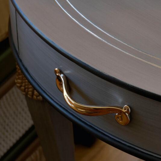 Exclusive Bedside Table With Tassel Design Detail Close-up image of an Exclusive Bedside Table With Tassel Design Detail featuring a sleek, curved, and polished gold-colored handle. The handle is slightly ornate with small curls at both ends, complementing the tassel design detail. It is mounted on a dark-stained surface with subtle grain patterns and a warm finish.