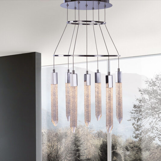 The Exclusive Chrome Pendant Light Cluster features a modern chandelier design with several elongated, textured glass pendants suspended from a sleek circular metallic base. Arranged beautifully in a tiered formation and supported by thin wires, this exclusive chrome fixture is set against a backdrop of large windows showcasing stunning views of trees and mountains.