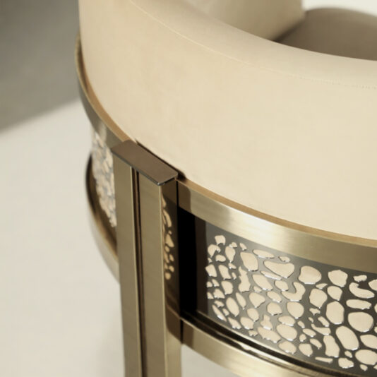 Exclusive Curved Bar Stool A close-up view of the Exclusive Curved Bar Stool, showcasing its beige seat and backrest. The metallic frame features an intricate, rock-like cutout pattern and boasts a modern, gold-colored finish.
