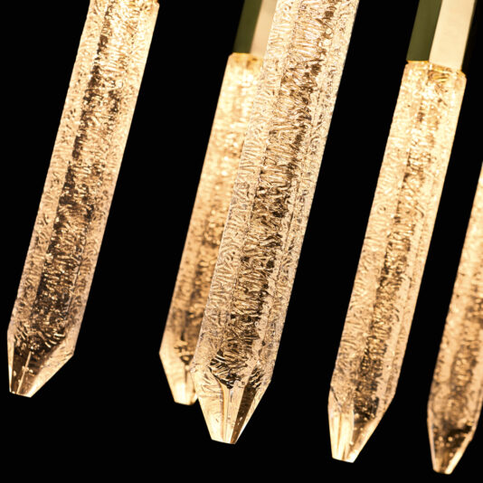 Exclusive Gold Vertical Crystal Chandelier Close-up of several illuminated, elongated glass light fixtures hanging against a black background. These fixtures taper to a pointed end, resembling stalactites, with an intricate bubble-like texture inside the glass that adds a sparkling effect, similar to the Exclusive Gold Vertical Crystal Chandelier.