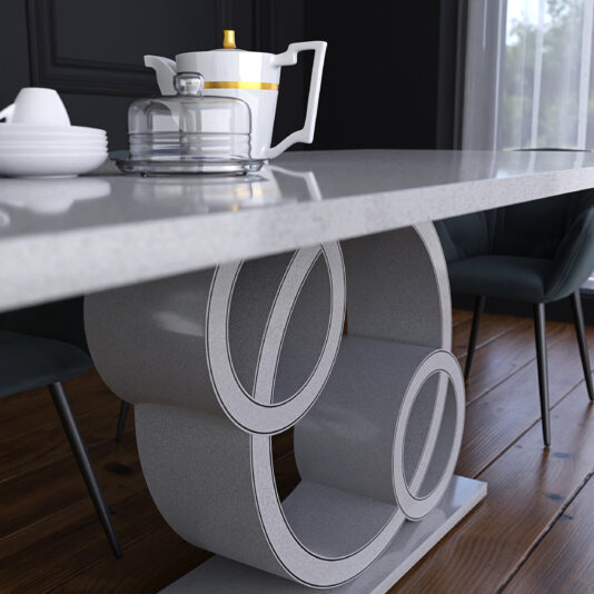 The Exclusive Marble Dining Table features a modern, unique curved base with circular cutouts. Atop the table is a white teapot accented with gold, a glass dome cake stand displaying a small cake, and a stack of white plates accompanied by a cup. Dark chairs and a window frame the background.