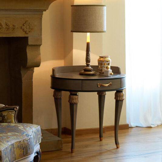 The Exclusive Side Table With Tassel Design Detail, featuring ornate legs and a drawer, sits next to a fireplace and a cushioned armchair. Adorned with a classic table lamp, the table also holds a decorative vase and a small cup, with sheer white curtains in the background.