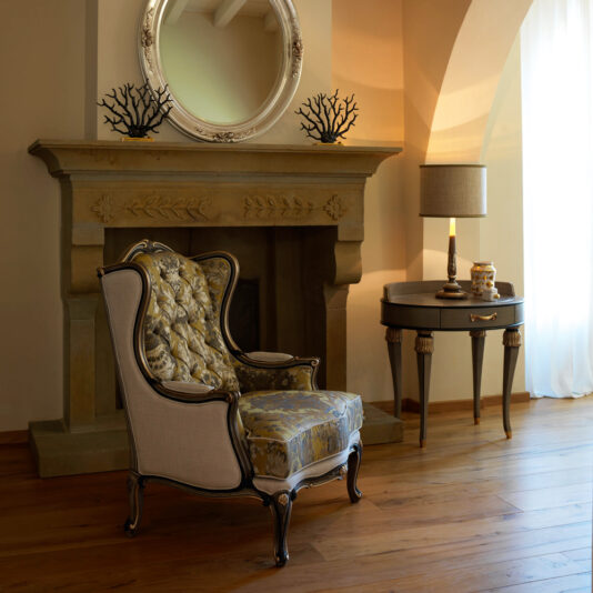 An elegant room with a vintage armchair featuring ornate upholstery sits in front of a stone fireplace. Above the fireplace, there is a decorative round mirror and black coral figurines. To the right, the Exclusive Side Table With Tassel Design Detail and ceramic decor sit near a sheer-curtained window.