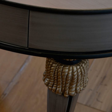 Exclusive Side Table With Tassel Design Detail Close-up of the Exclusive Side Table With Tassel Design Detail, showcasing its light grey finish. The edge features a smooth curve complemented by intricately carved, gold-colored detailing on the leg that mimics scaled design patterns. Tassel design details add a unique touch, all beautifully set against a polished wooden floor.