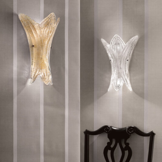 Two elegant, sculptural Glass Leaf Wall Lights are mounted on a patterned wall with vertical gray and beige stripes. These Glass Leaf Wall Lights feature a wavy, leaf-like design, one emitting a warm light and the other a cool light. The back of a dark wooden chair is partially visible at the bottom.