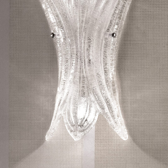The Glass Leaf Wall Light, featuring an elegant, textured glass leaf and a modern, curved design, is mounted against a grid-patterned wallpaper. The light shines through the glass, highlighting its intricate details and casting subtle shadows on the wall.
