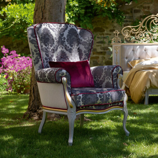 A High Backed Venetian Style Winged Armchair, showcasing a floral pattern with purple accents, is placed on a lush green lawn beside a tree. In the background, an ornate bed adds to the scene's charm as bright sunlight enhances the garden's vibrant colors.