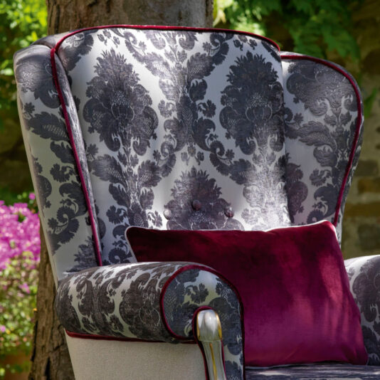 The High Backed Venetian Style Winged Armchair, adorned with luxurious ornate dark gray damask upholstery, stands elegantly outdoors. The chair is accented with purple velvet piping and accompanied by a deep burgundy velvet pillow. A serene garden setting is revealed in the background, showcasing bright flowering plants and a tree trunk.
