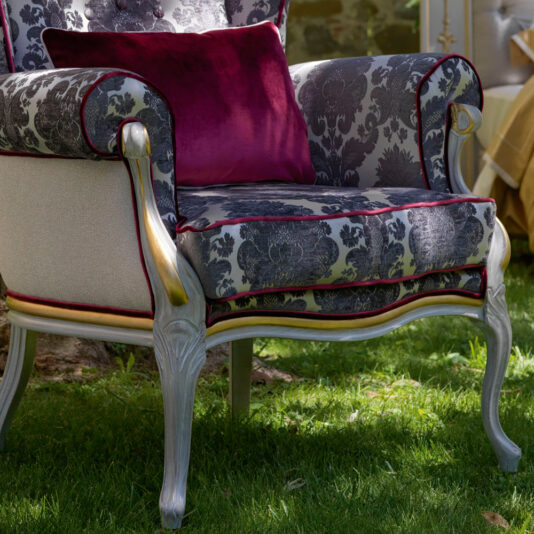 An elegant High Backed Venetian Style Winged Armchair features a sophisticated floral damask pattern in grayscale, accented by red piping and silver-toned wood. It is complemented by a deep burgundy throw pillow and positioned gracefully on a grassy lawn in an outdoor setting.