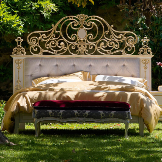 In a lush garden setting, a High End Venetian Style Brass Bed with an ornate, gold-trimmed frame and an intricate headboard is elegantly displayed. The bed features cream-colored bedding topped with a mustard yellow duvet, complemented by a small bench upholstered in velvet with a dark floral pattern at its foot.