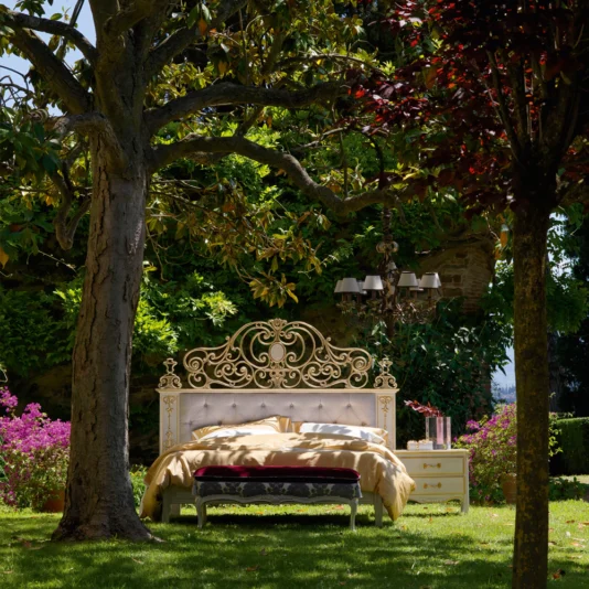A luxurious High End Venetian Style Brass Bed with an ornate headboard is set outdoors in a lush garden under a large tree, surrounded by vibrant pink flowers and greenery. The serene setting is enhanced by a nearby chandelier.