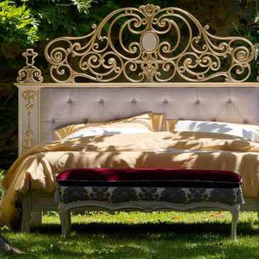 High End Venetian Style Brass Bed The High End Venetian Style Brass Bed, adorned with an ornate gold-painted headboard featuring intricate swirling designs and topped with a small decorative urn, exudes elegance. Dressed in cream and beige bedding, it is complemented by a red and black upholstered bench at its foot and surrounded by lush greenery.