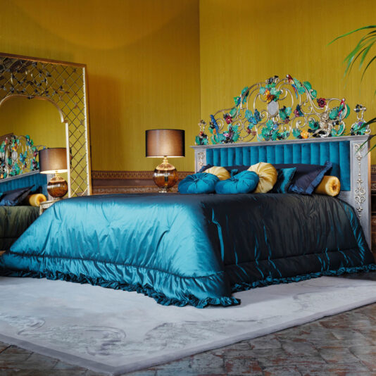 A luxurious, tropical-themed bedroom features a plush turquoise bed with an ornate, multicolored headboard adorned with intricate designs. This High End Venetian Style Tropical Brass Bed is flanked by two bedside tables, each with a gold lamp sporting a black shade. The room's yellow walls complement a large, decorative mirror.