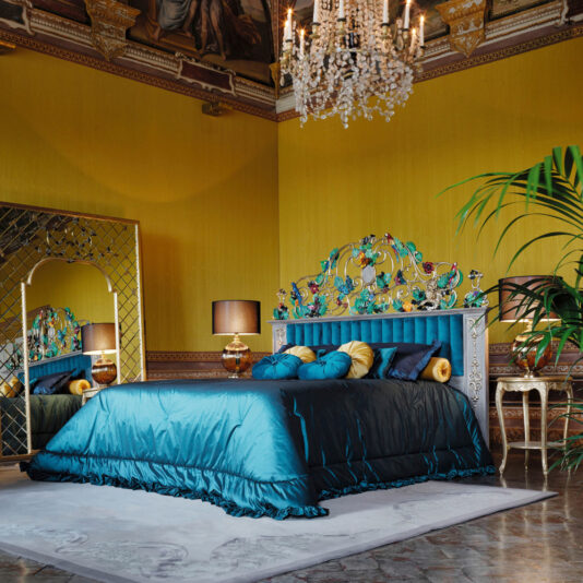 High End Venetian Style Tropical Brass Bed Luxurious bedroom featuring an ornate, colorful headboard and the High End Venetian Style Tropical Brass Bed with teal bedding and decorative pillows. A large mirror, chandelier, and elegant furnishings enhance the opulent yellow-gold decor, while a potted plant adds greenery.