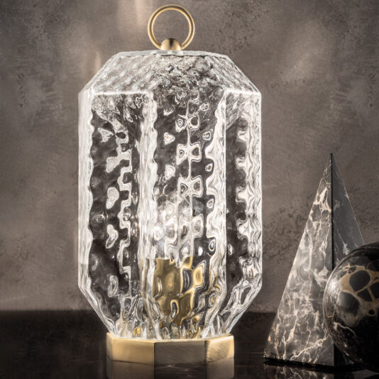 The Lantern Style Murano Glass Lamp, featuring a hexagonal glass design with a brass base and handle, is placed on a reflective dark surface alongside a black marble pyramid and sphere. The background is set against a grey textured wall.