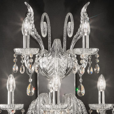 A close-up image of an ornate crystal chandelier showcasing multiple candle-shaped light bulbs and hanging crystal droplets, set against a dark background. The intricate detailing and reflective surfaces create a luminous and elegant appearance, reminiscent of the Large Acanthus Glass Wall Light.