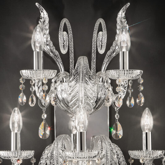A close-up image of an ornate crystal chandelier showcasing multiple candle-shaped light bulbs and hanging crystal droplets, set against a dark background. The intricate detailing and reflective surfaces create a luminous and elegant appearance, reminiscent of the Large Acanthus Glass Wall Light.