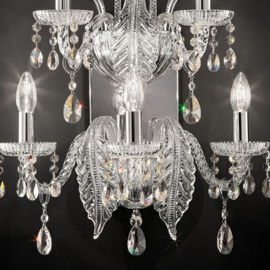 A close-up of the Large Acanthus Glass Wall Light reveals an ornate chandelier with five candle-shaped light bulbs. This exquisite fixture boasts intricate glass designs and numerous dangling crystal prisms, creating a sparkling effect against a dark background.