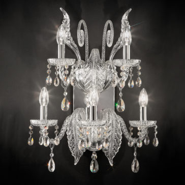 An ornate crystal chandelier with five candle-shaped light bulbs, intricately designed crystal arms, and numerous hanging teardrop-shaped prisms stands beside the Large Acanthus Glass Wall Light, all set against a dark background, creating a luxurious and elegant appearance.