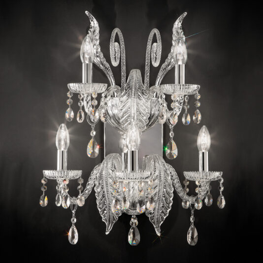 An ornate crystal chandelier with five candle-shaped light bulbs, intricately designed crystal arms, and numerous hanging teardrop-shaped prisms stands beside the Large Acanthus Glass Wall Light, all set against a dark background, creating a luxurious and elegant appearance.