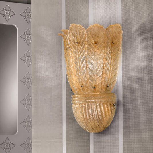 The Large Amber Glass Leaf Wall Light, featuring a golden, textured shade that is artfully designed to resemble an open flower or leaves, is mounted against neutral-striped wallpaper. To the left of the fixture, a portion of an ornate mirror can be seen.