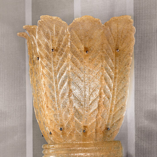 Large Amber Glass Leaf Wall Light A close-up of the Large Amber Glass Leaf Wall Light, an ornate and golden fixture, mounted on a textured wallpaper featuring vertical striped patterns. The intricately designed light gives the appearance of overlapping, intricately detailed leaves.