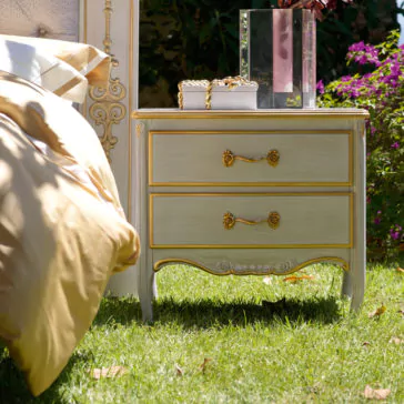 A Large Classic Two Drawer Bedside Cabinet, featuring gold accents and ornate handles, stands on a grassy lawn next to a bed adorned with golden sheets. On the nightstand are a white decorative box and a tall vase of flowers, set against green foliage and purple blooms in the background.