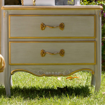 A large Classic Two Drawer Bedside Cabinet with ornate golden handles and decorative carvings stands on a grassy surface. A small cat is partially visible lying underneath, blending into the shadow.