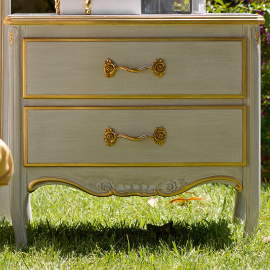 A large Classic Two Drawer Bedside Cabinet with ornate golden handles and decorative carvings stands on a grassy surface. A small cat is partially visible lying underneath, blending into the shadow.