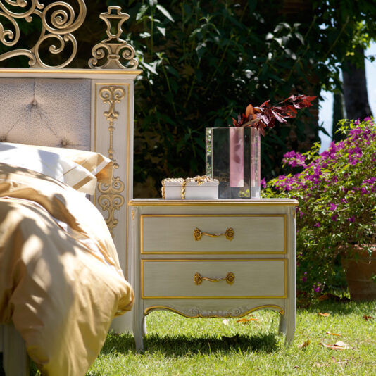 An opulent outdoor bedroom arrangement includes a baroque-style bed adorned with an intricate headboard and golden bedding. Beside the bed stands a Large Classic Two Drawer Bedside Cabinet, topped with a glass vase filled with red foliage and small decorative items, all set amidst abundant greenery and blooming flowers.