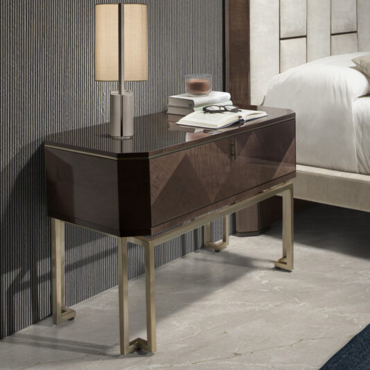 A Large Dark Maple Bedside Table boasts a glossy wooden surface and metallic legs. Its top is adorned with a table lamp, an open book, glasses, and a small glass candle holder. The background features a vertical striped wall and part of a bed with a tufted headboard.