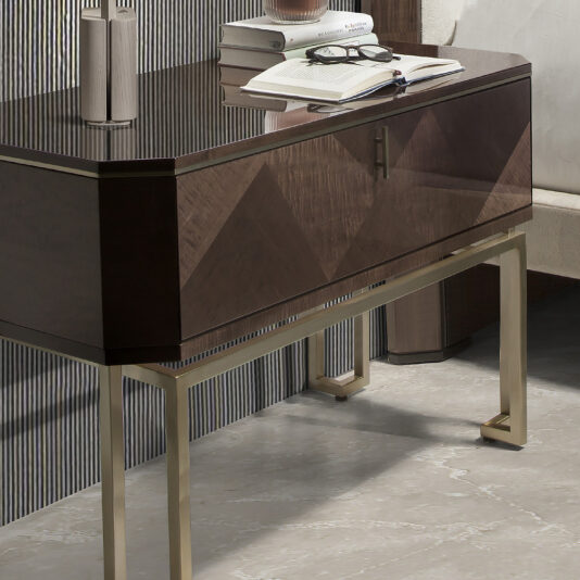 Large Dark Maple Bedside Table A sleek modern desk with a glossy wooden surface and metal legs, showcasing an open book, stacked closed books, and a pair of eyeglasses on top. The background features a striped wall, and to the right, partially visible is a fabric-covered chair paired with the Large Dark Maple Bedside Table.