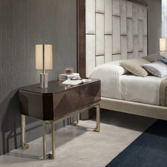 Large Dark Maple Bedside Table A modern bedroom features a sleek Large Dark Maple Bedside Table with gold-colored legs, holding a beige lamp and a couple of books. The large bed has a cushioned, geometrically patterned headboard, and beige and white bedding. The room is elegant with neutral tones.