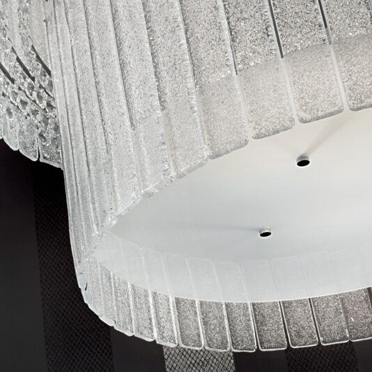 Large Modern Tiered Glass Chandelier A Large Modern Tiered Glass Chandelier from hangs from the ceiling, composed of multiple horizontal layers of rectangular, textured glass panels that create a cascading effect. The chandelier’s central part is white and smooth, complemented by a dark contrasting wall in the background.