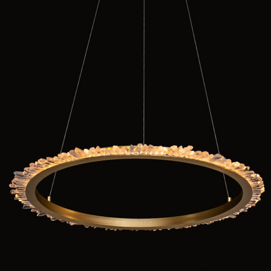 Large Quartz Crystal Gold Round Chandelier The Large Quartz Crystal Gold Round Chandelier hangs suspended by thin wires, its golden frame adorned with small quartz crystal embellishments. It emits a warm, ambient glow against a black background.