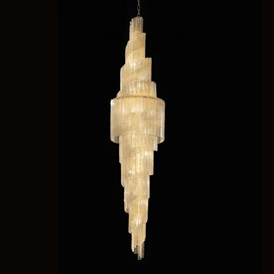 Large Tiered Amber Glass Chandelier The Large Tiered Amber Glass Chandelier is an elegant fixture composed of numerous vertical tubes with a tiered, cascading design. It emits a warm, golden glow and its intricate and luxurious structure is accentuated against a black background.