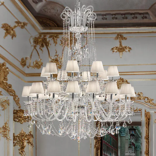 A Large Two Tier Crystal Chandelier With Shades, featuring white lampshades, hangs from an elaborately decorated ceiling in a luxurious room adorned with gold accents and intricate wall designs. The background showcases detailed murals and decorative molding.