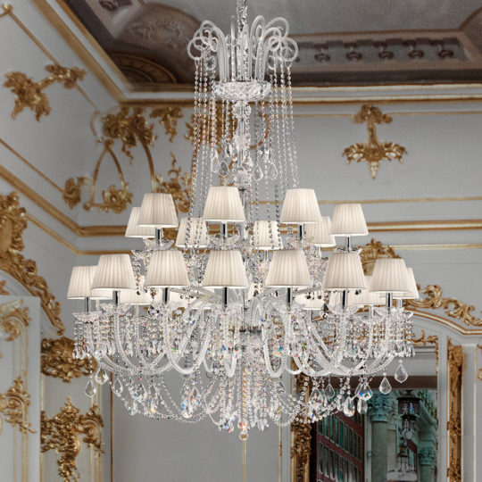 A Large Two Tier Crystal Chandelier With Shades, featuring white lampshades, hangs from an elaborately decorated ceiling in a luxurious room adorned with gold accents and intricate wall designs. The background showcases detailed murals and decorative molding.