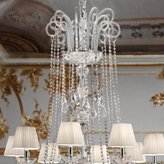 The Large Two Tier Crystal Chandelier With Shades, featuring cascading strands of beads and intricate designs, hangs from an ornate ceiling. Below the crystals, elegant lampshades emit a warm light. The ceiling's detailed golden patterns and light brown accents further enhance the chandelier's opulence.