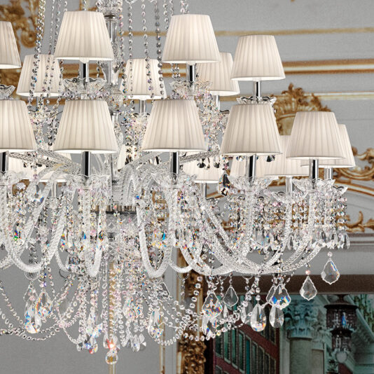 The Large Two-Tier Crystal Chandelier With Shades is an ornate fixture adorned with crystal beads and hanging pendants, each arm crowned with a small lampshade. In the background, an elegant room showcases intricate gold detailing and a beautifully painted ceiling.