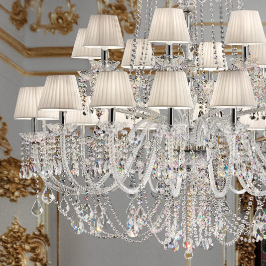 The Large Two Tier Crystal Chandelier With Shades is adorned with multiple intricate hanging crystals and elegant lampshades. Set against an ornately decorated background with gold accents, it evokes a sense of opulence and grandeur.