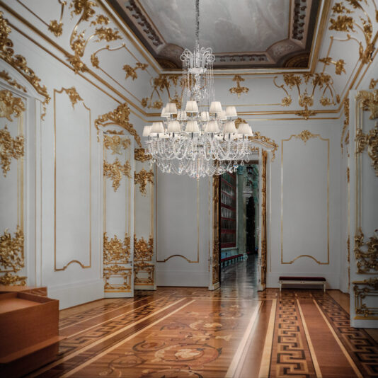 An ornate room adorned with white and gold walls, intricate moldings, and a **Large Two Tier Crystal Chandelier With Shades**. The wooden floor showcases detailed inlays. An open doorway at the back reveals a hallway lined with bookshelves. The ceiling is embellished with decorative paintings.