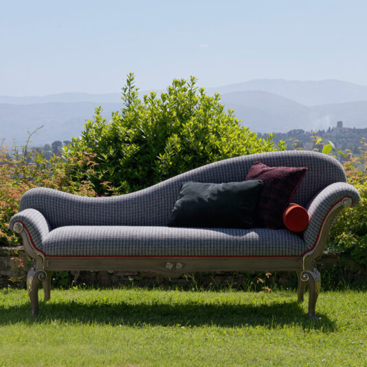A stylish Louis Style Classic Chaise Longue with dark blue upholstery is placed outside on a well-manicured lawn. The chaise features two cushions, one black and one red, and is situated in front of a lush green hedge with expansive mountains in the distance under a clear sky.