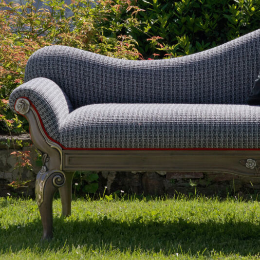 The ornate Louis Style Classic Chaise Longue, featuring wooden legs, is set on a lush green lawn with leafy bushes in the background, adding a touch of classic elegance to the outdoor setting.