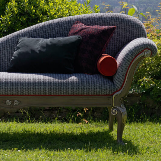 The Louis Style Classic Chaise Longue, featuring ornate wooden legs and grey upholstery with red trim, is elegantly placed on a lush green lawn. It is complemented by a deep red throw pillow and a black cushion, with verdant foliage and rolling hills in the background.