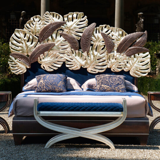 The Luxurious Bed With Palm Leaf Design Headboard features a beautiful ornate design with large metallic leaves. Complemented by patterned blue and beige linens and a curved wooden bench at the foot, it creates an elegant atmosphere amid lush greenery.
