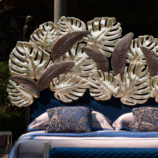 Luxurious Bed With Palm Leaf Design Headboard The Luxurious Bed With Palm Leaf Design Headboard features large metallic leaves in silver and bronze tones. Adorned with navy blue and white pillows and a matching navy blue blanket, the setting appears to be outdoors, with greenery and statues partially visible in the background.
