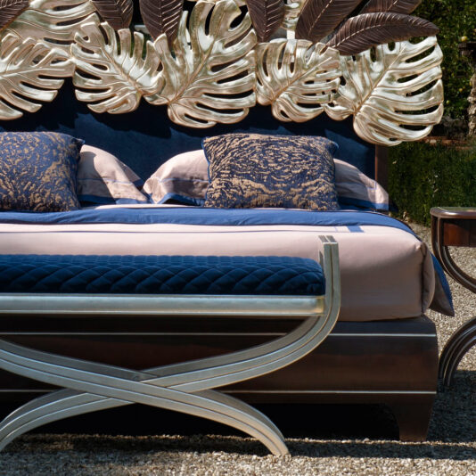 Luxurious Bed With Palm Leaf Design Headboard The Luxurious Bed With Palm Leaf Design Headboard showcases large metallic leaves, paired with a dark blue bedspread and abstract-design pillows. An ornate bench with a metallic finish is positioned at the foot of the bed, creating an outdoor ambiance.