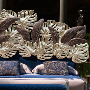 Luxurious Bed With Palm Leaf Design Headboard The Luxurious Bed With Palm Leaf Design Headboard showcases an elaborate arrangement of large metallic tropical leaves. Adorned with blue and beige bedding, complemented by blue patterned cushions, this bed is set in an elegant outdoor area surrounded by classical statues and lush greenery.